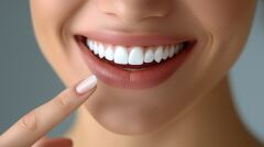 A woman beams with joy, pointing at her flawless white teeth against a gray backdrop. Her confident smile highlights the importance of dental health and care.