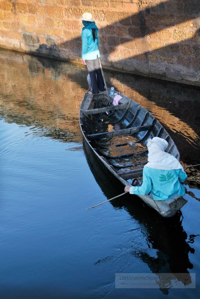 Two individuals navigate a tranquil waterway in Siem Reap Cambodia surrounded by historic architecture. The calm blue waters reflect the beauty of the ancient environment at sunset creating a peaceful ambiance.