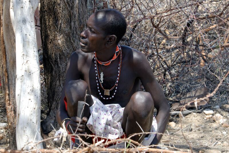 A Samburu man wearing traditional beads and attire sits by a tree in the arid landscape of Kenya focused on crafting The surrounding environment showcases the natural beauty and cultural heritage of the Samburu community