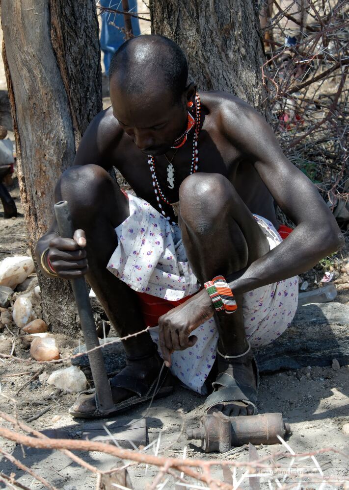 A Samburu man sits on the ground skillfully using traditional tools to craft an item He is dressed in traditional attire surrounded by the natural landscape of Samburu in Kenya showcasing the cultural craftsmanship of the tribe