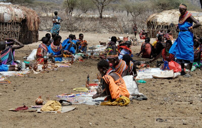 Members of the Samburu tribe in Kenya gather on the ground to create and sell traditional crafts Vividly dressed in colorful clothing they display their handiwork while surrounded by simple structures and the arid landscape typical of the region