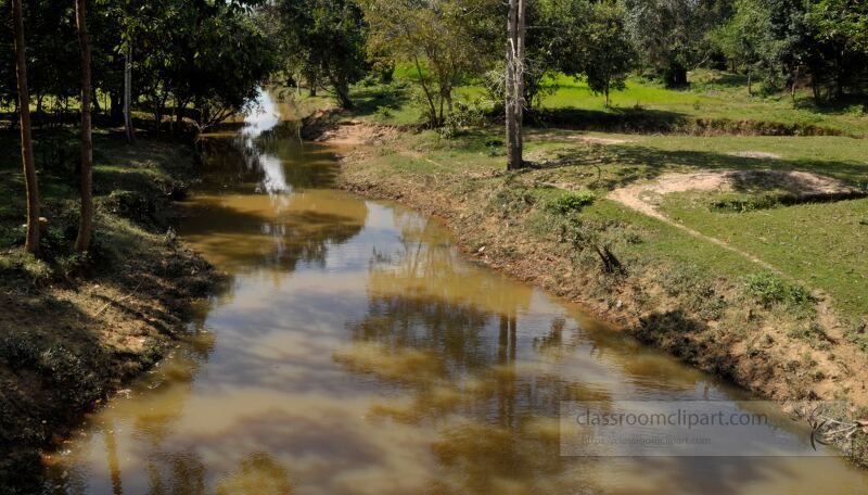 Tranquil waters reflect a clear sky as lush greenery frames a peaceful riverside in Siem Reap Cambodia. Nature thrives in harmony inviting visitors to admire the beauty and serenity of the surroundings.